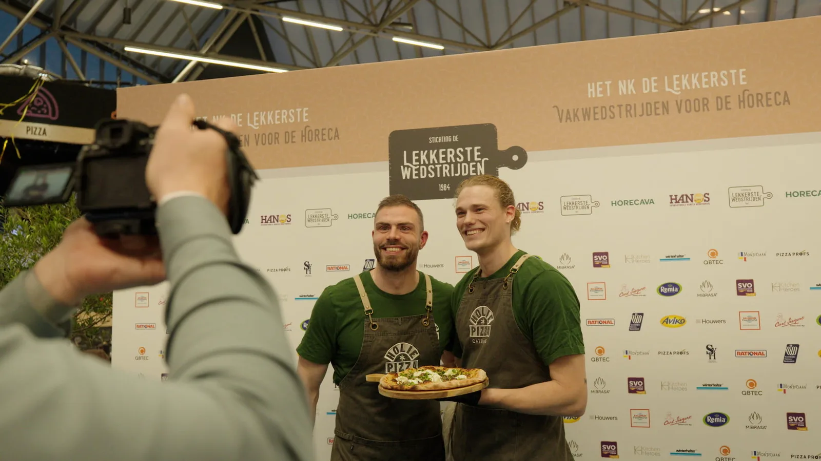 Twee pizzabakkers van Holie Pizza poseren met hun pizza voor de perswand van het NK De Lekkerste