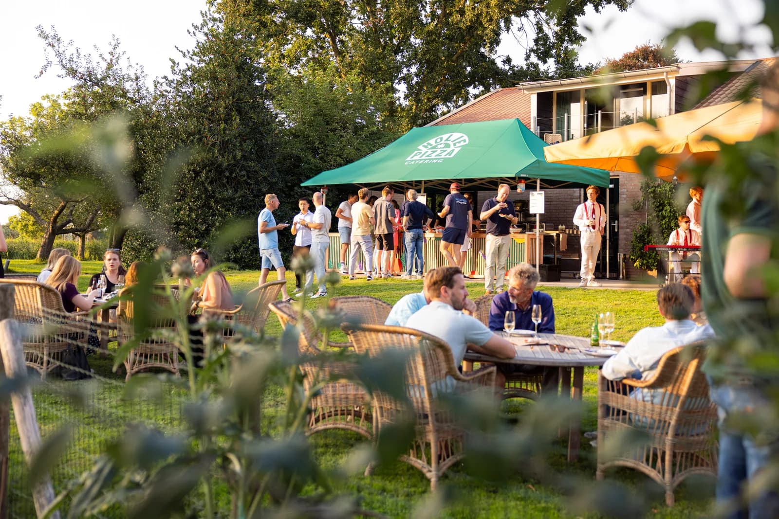 Gasten aan tafel in een tuin, met de mobiele pizzakeuken van Holie Pizza onder de groene tent